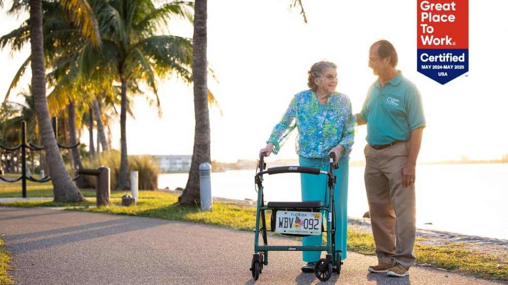 Senior woman using a walker and Right at Home male caregiver on a sidewalk with a Great Place to Work 2024 badge in the upper right corner