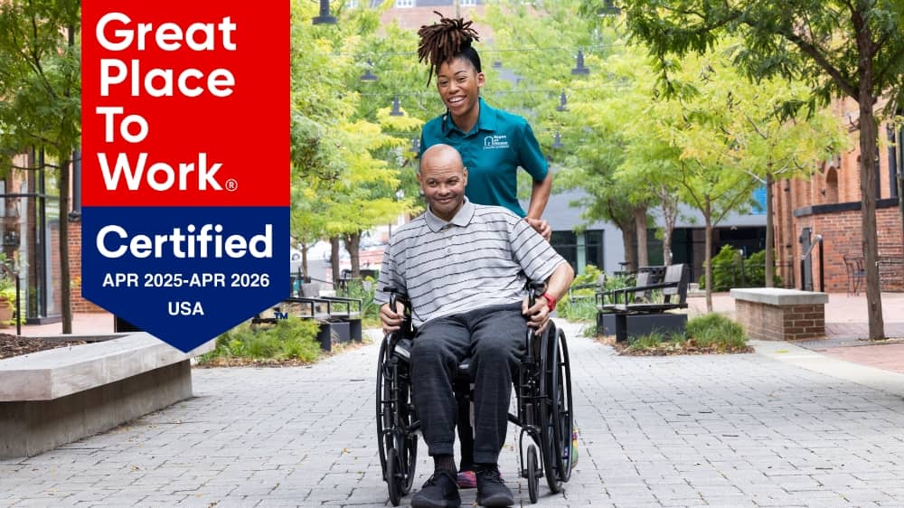 Female Right at Home caregiver pushing a man in a wheelchair with a 2025 Great Place to Work certification badge on the left side of the photo.