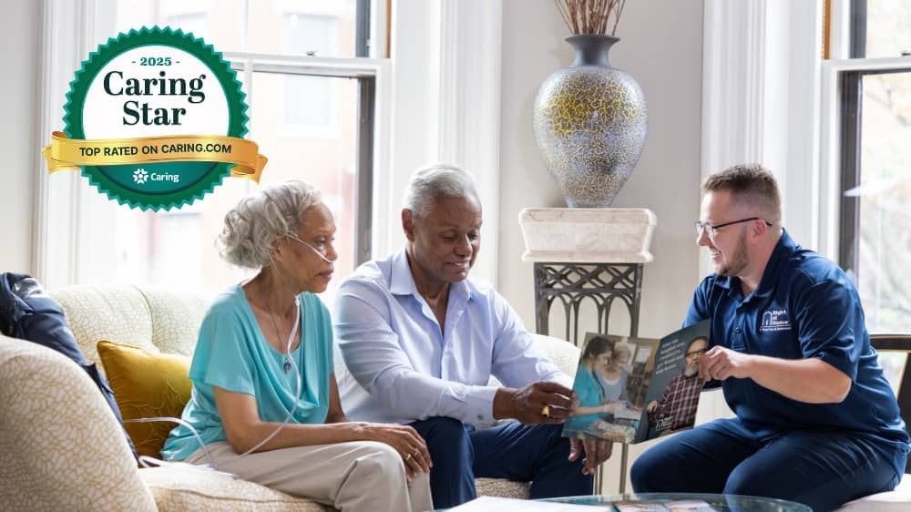 Senior male and female couple sitting on a couch across from a Right at Home caregiver looking at a care phamplet with a 2025 Caring Star award badge in the upper left corner of the image