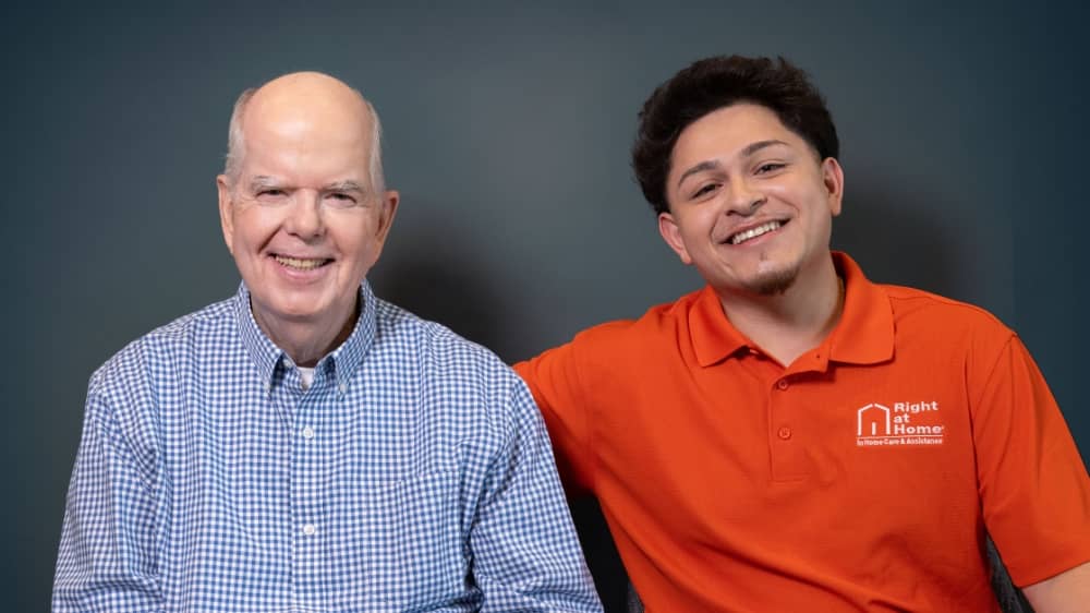 Senior male client sitting next to a male Right at Home caregiver against a dark grey background
