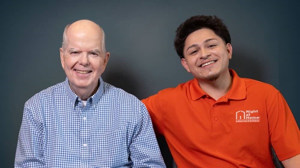 Senior male client sitting next to a male Right at Home caregiver against a dark grey background