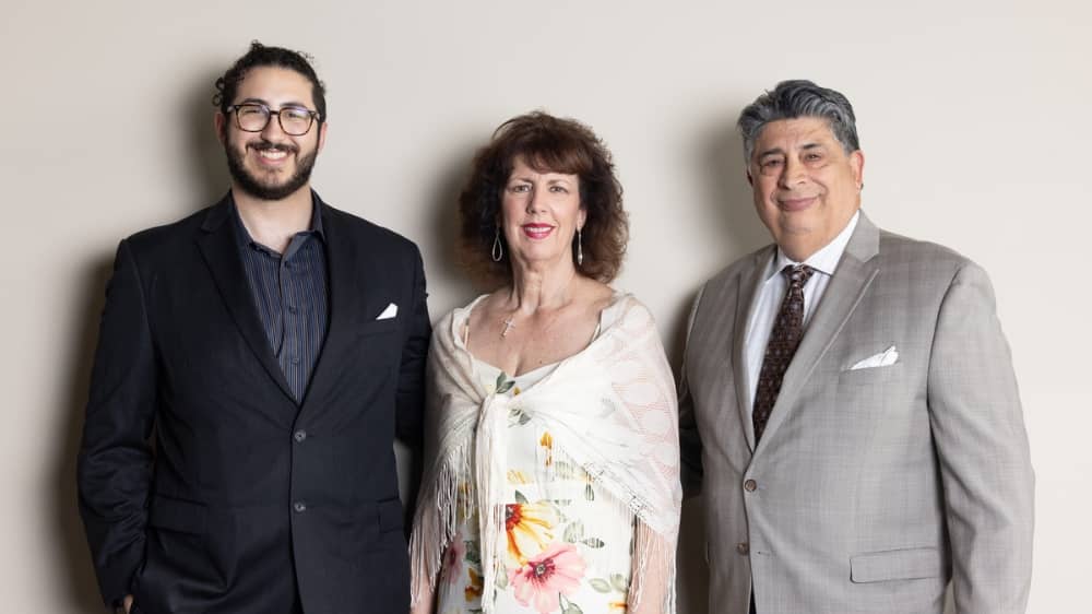 Concialdi family, father, mother, and adult son, smiling and standing against a plan white background