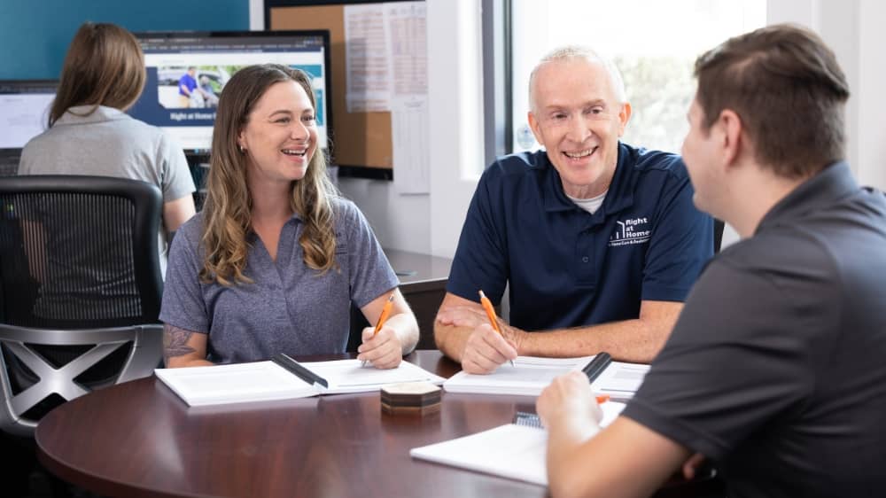 One woman and two men sitting at an office table with notebooks in front of them talking and laughing 