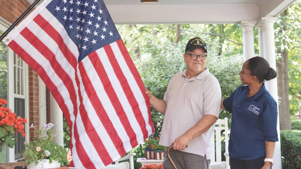 Senior male veteran standing with a female Right at Home Caregiver. Both of them are looking at an American flag which is mounted on the porch