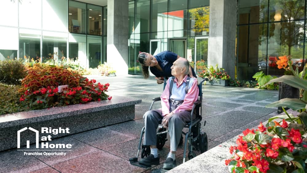 Female Right at Home caregiver pushing a male senior client in a wheelchair outside of a large building