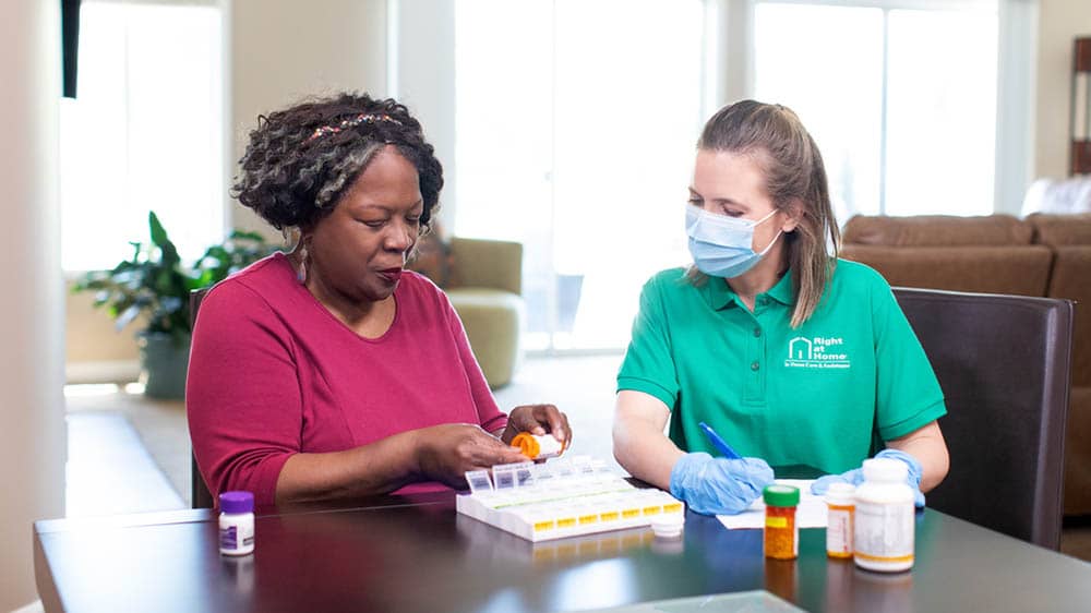 A female Right at Home caregiver helping monitor a senior female client sort medications for the week