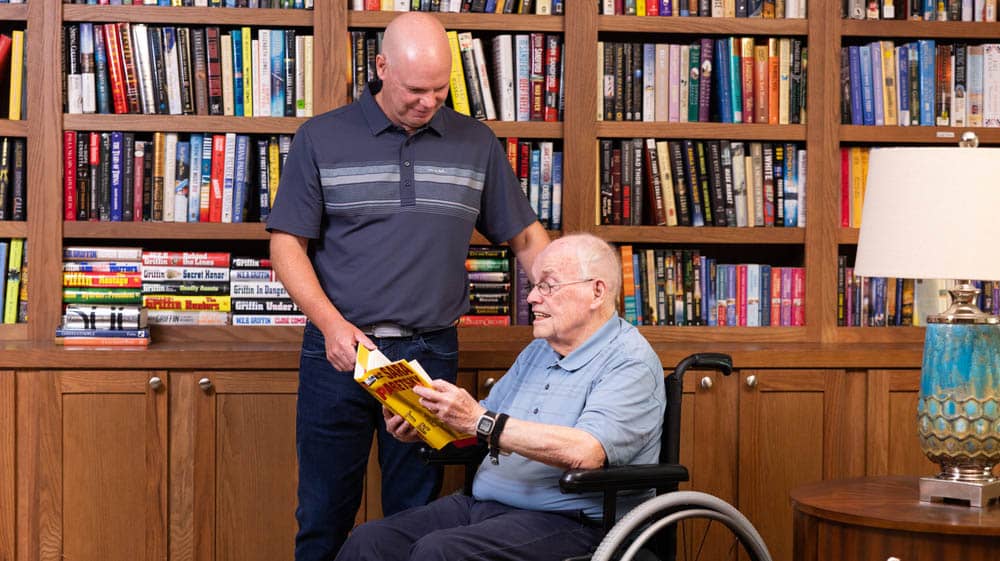 A male Right at Home caregiver assisting a senior male client in a wheelchair pick out a book in a home library 
