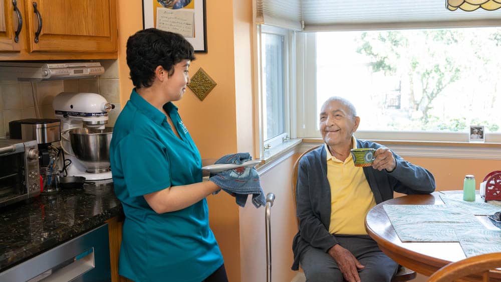 A female Right at Home caregiver is drying a dish while her senior male client sits at the kitchen table enjoying a coffee