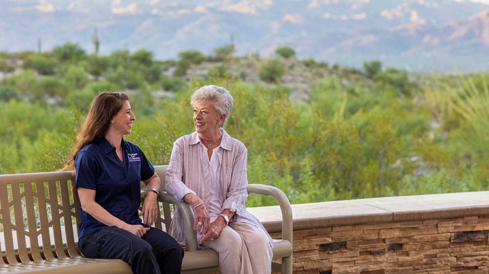 A female Right at Home caregiver is sitting on a bench outside with her female senior client