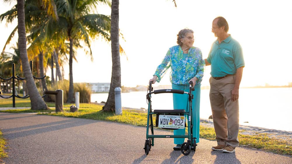 A male Right at Home caregiver is standing next to his female senior client using a walker outside next to a body of water