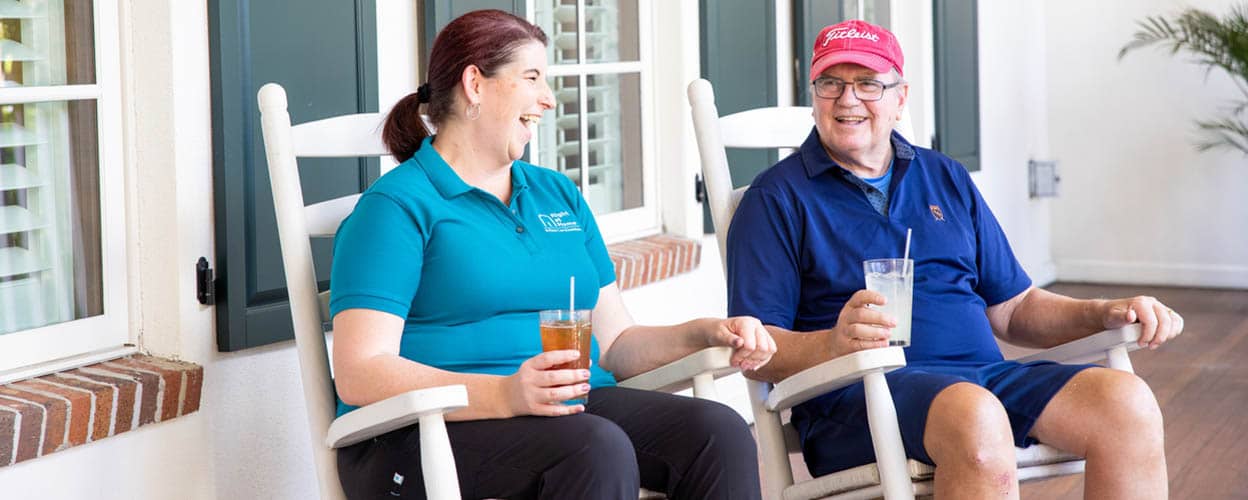 Female Right at Home caregiver and her senior male client are sitting in rocking chairs on a porch enjoying a cold beverage