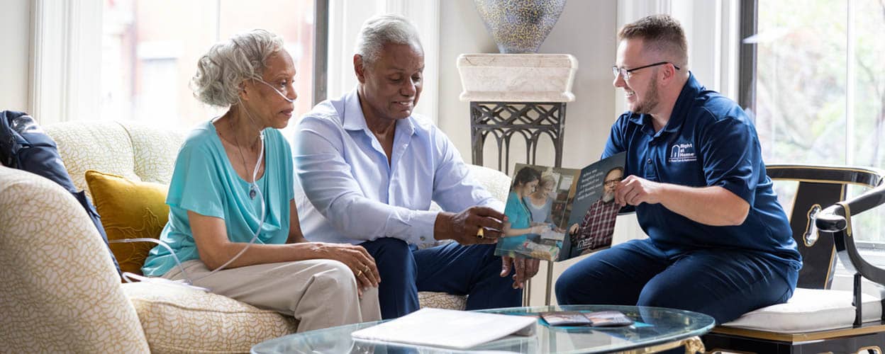 A male Right at Home representative is meeting with a senior couple in their living room for a consultation 