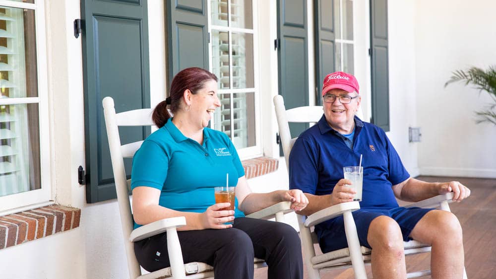 A senior male client sitting in a rocking chair with a cold drink next to a female Right at Home caregiver sitting in a rocking chair on the front porch