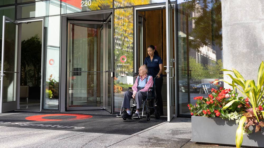 A female Right at Home caregiver is pushing her senior male client in his wheelchair out the front door of a medical facility