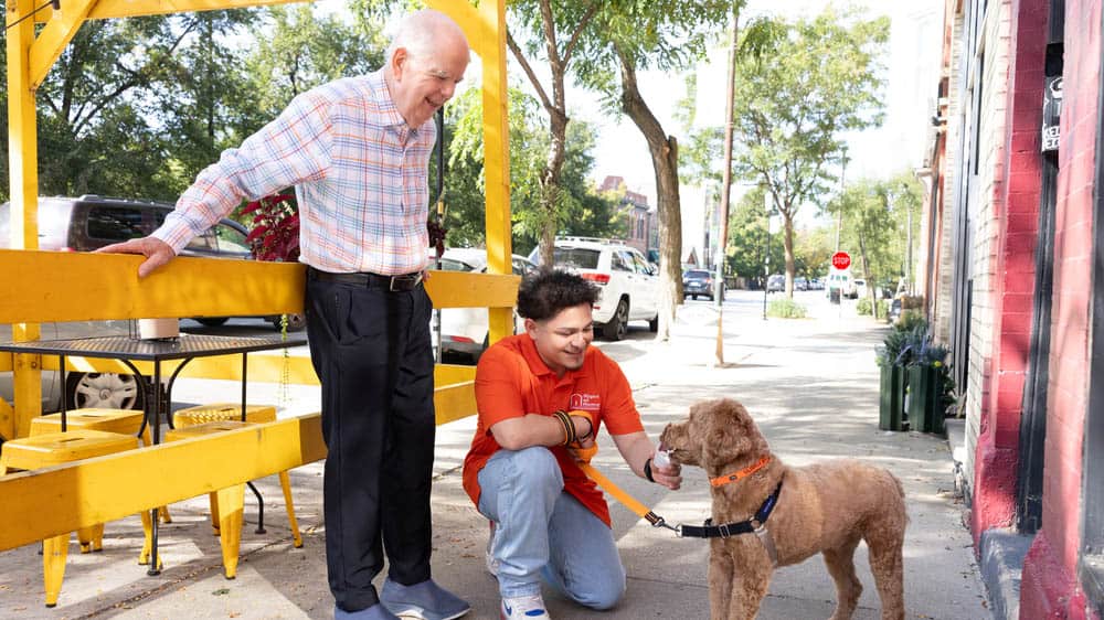 A senior male client looks on as his male Right at Home caregiver is crouched down feeding a dog a pup-cup while walking outside