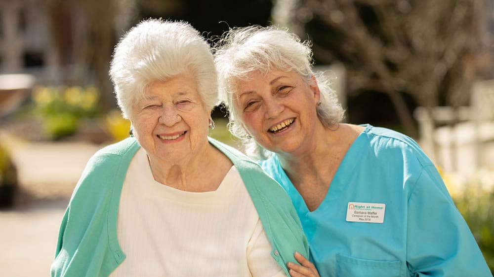 A female senior client and female caregiver smiling while standing together outside