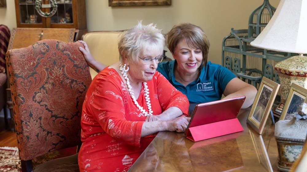 A female senior Right at Home client is looking at her tablet set up at her desk while her female Right at Home caregiver is helping point something out to her