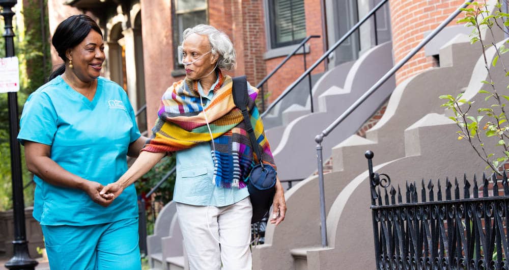 A senior female wearing an oxygen tube, is walking hand in hand with a female Right at Home caregiver outside.